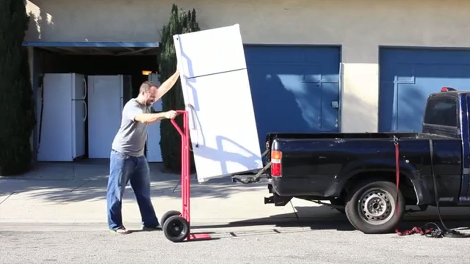 a mover loads a white refrigerator onto a pickup truck with a hand truck on a sunny street
