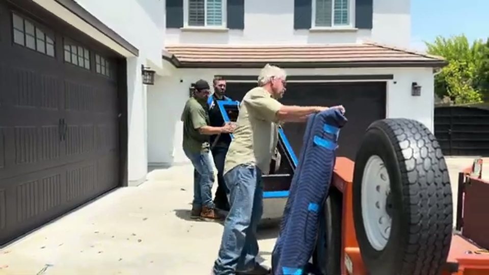 three-man crew unloading a blanket-wrapped safe from a flatbed trailer in a driveway, face visible